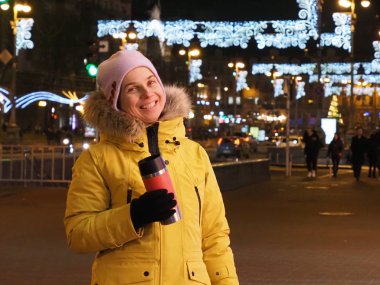 Soğuk mevsimde bir gece şehrinin arka planında gülümseyen Ukraynalı kadın sıcak bir bardaktan sıcak çay içerek çelenk yakıyor.