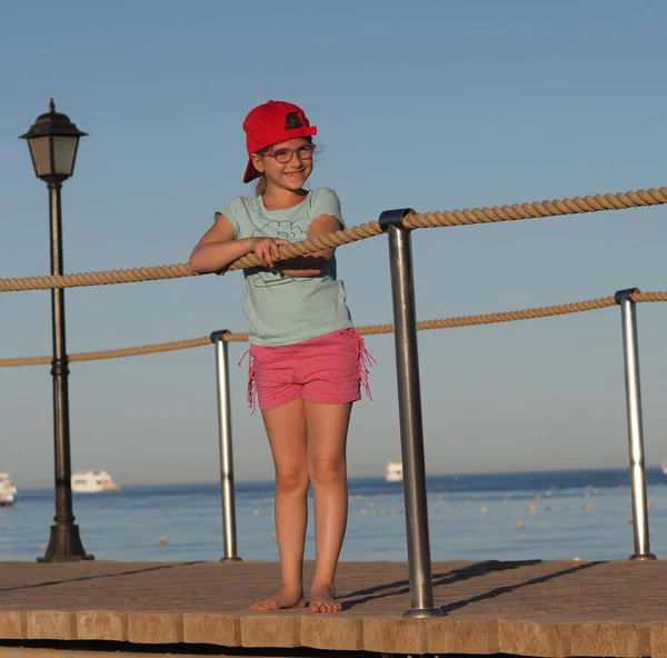 船や夕方の青空を背景に 海を背景に提灯を持つ木製の桟橋に赤いキャップの女の子 — ストック写真