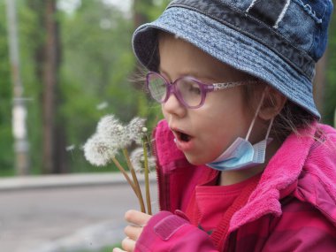 Karantina sırasında gözlüklü, kot şapkalı ve tıbbi maskeli Avrupalı bir kız karahindiba tohumları saçar.