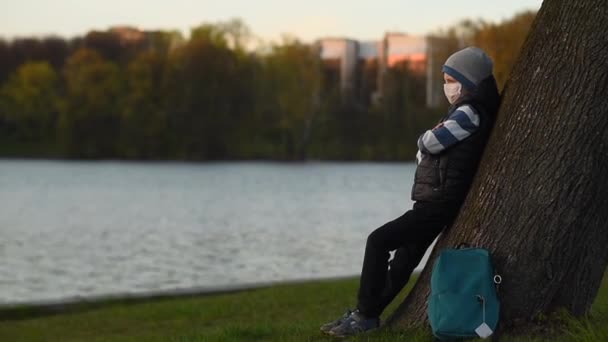 En ung människa i medicinsk mask väntar på att luta sig mot ett träd på flodstranden och lämnar — Stockvideo