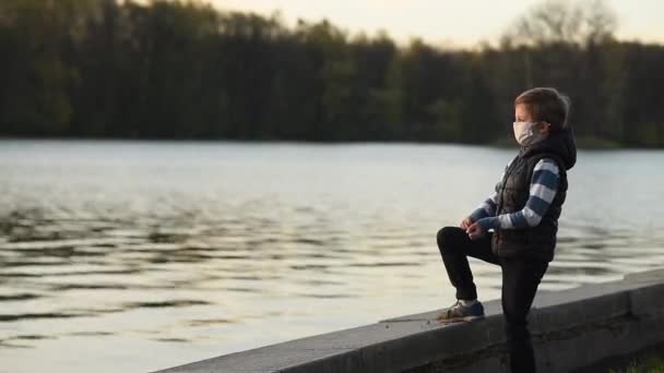 Un ragazzo con una maschera medica sulla riva getta pietre in acqua al tramonto — Video Stock