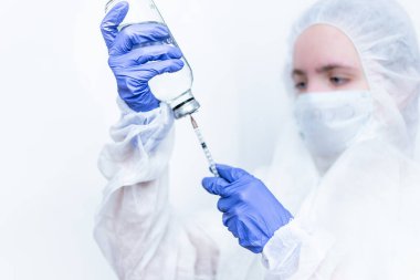Virüs geçirmez giysi ve tıbbi maske giyen bir virüs uzmanı şırıngayı pembe ilaçla dolduruyor. Beyaz bir arka planda elinde şırıngayla kadın bir hemşire..