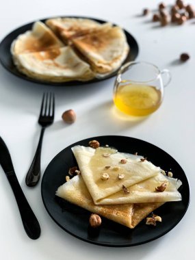 Beyaz tabakta, siyah tabakta bal ve fındıklı krepler. Kahvaltı ya da tatlı için ev yapımı ince krepler..