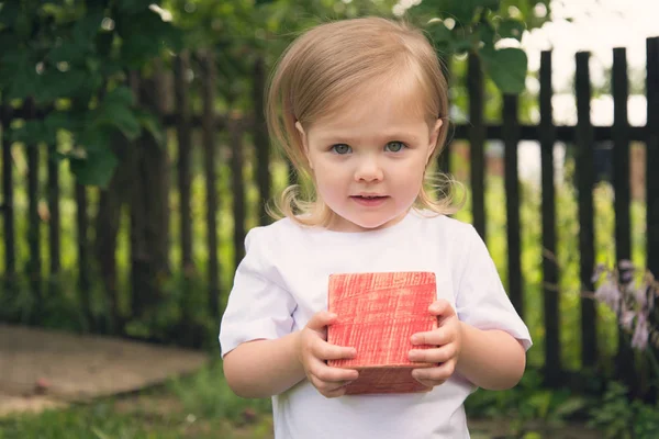 Bahçede beyaz tişörtlü bir kız elinde kırmızı tahta bir küp tutuyor.