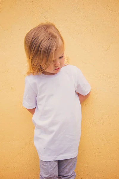 Üç yaşında beyaz tişörtlü bir kız sarı duvarlı bir modelin karşısında.