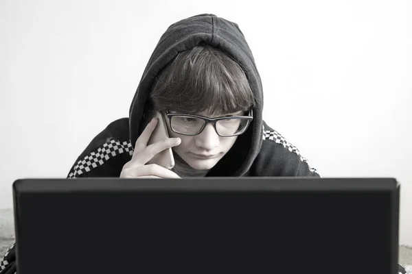 Мужчина в черном капюшоне смотрит на ноутбук и разговаривает по телефону — стоковое фото