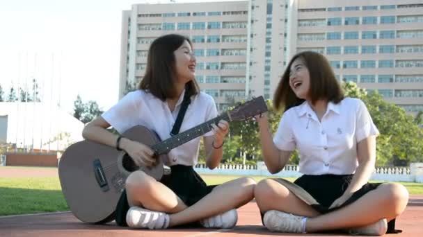 Χαριτωμένο ασιατικό Ταϊλάνδης Κολέγιο Μαθητριεσ Ζευγάρι κορίτσι σε Πανεπιστήμιο ομοιόμορφη sit τραγουδώντας και παίζοντας κιθάρα έχοντας διασκεδαστικό υπαίθριο με ευτυχισμένη έκφραση. Σε φιλικό μαθήτρια φοιτητής κορίτσι αναψυχής έννοια. — Αρχείο Βίντεο