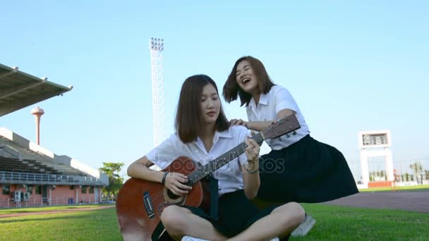 Lindo asiático tailandés estudiante universitario chica en uniforme universitario tocando la guitarra y otra chica ven y abrazarla en la espalda en la amistad amorosa colegiala y estudiante universitaria chica concepto de ocio en HD . — Vídeos de Stock