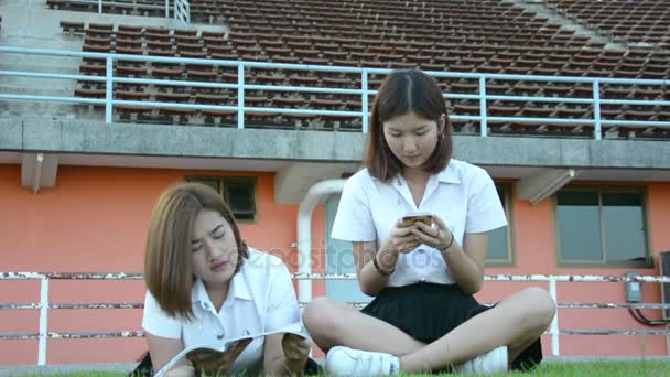 Niedliche asiatische thailändische College-Studentin Mädchen in Uniform der Universität Textbuch lesen und fragen Sie ihre Freundin, dass das Spielen mit dem Handy zu erklären, was sie nicht verstehen, in entspannendes Studienkonzept in hd — Stockvideo
