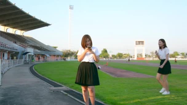 Cute Asian Thai college student girl in university uniform playing with her mobile and another girl come and close her eyes with surprise and hugging each other in loving friendship leisure concept. — Stock Video