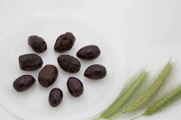 Pocas aceitunas negras oscuras en un plato blanco y tres espigas verdes o — Foto de Stock