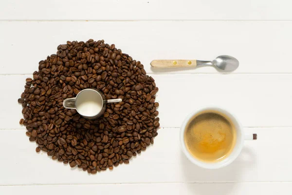 Pitcher with milk in the pile of coffee beans on the white wooden background. Spoon, mug and  white cup with coffee . Food and drink background photography.