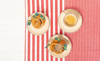 İsveç tarçınlı çörekler (kanelbullar) ve beyaz ahşap masada kahve. İsveç bayrakları. Çizgili masa örtüsü. Kahve molası (fika) kavramı. Üst görünüm. 