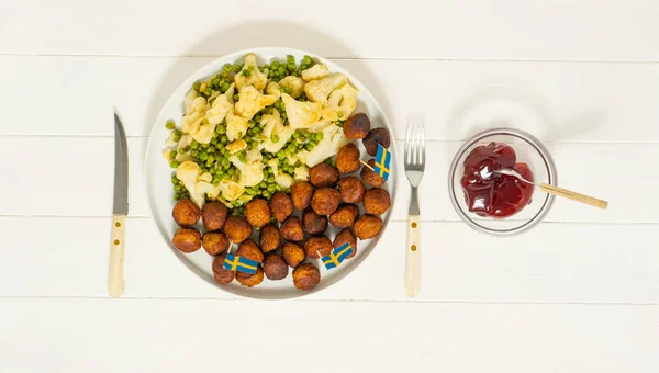 Traditional swedish meatballs (kottbullar) with cauliflower and green peas, lingonberry sauce in the white plate. Swedish flags. Swedish national food concept. Background photo. Top view.