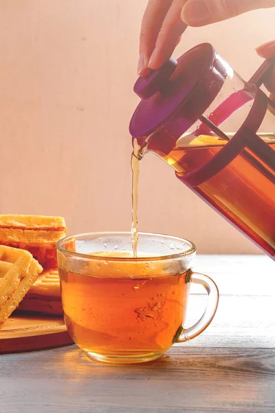 Female Hands Pour Tea from Brewer into Mug on White Wooden Table with Waffles. — Stock Photo, Image
