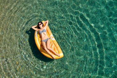Bikinili sıska genç bir kadın ve şişme yatakta güneş gözlüğü açık denizde yüzüyor..