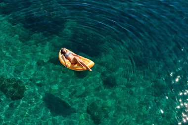 Bikinili sıska genç bir kadın ve şişme yatakta güneş gözlüğü açık denizde yüzüyor..