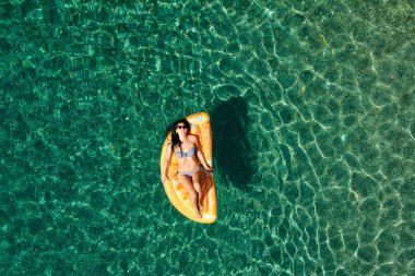 Bikinili sıska genç bir kadın ve şişme yatakta güneş gözlüğü açık denizde yüzüyor..