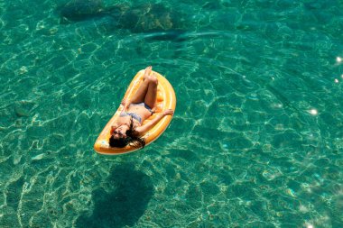Bikinili sıska genç bir kadın ve şişme yatakta güneş gözlüğü açık denizde yüzüyor..