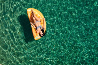 Bikinili sıska genç bir kadın ve şişme yatakta güneş gözlüğü açık denizde yüzüyor..
