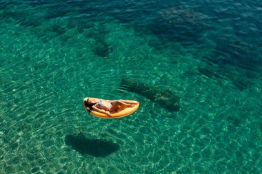 Bikinili sıska genç bir kadın ve şişme yatakta güneş gözlüğü açık denizde yüzüyor..
