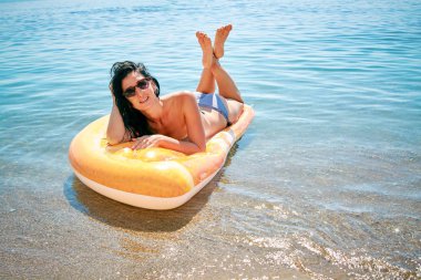 Bikini ve güneş gözlüklü sıska genç kadın deniz kenarındaki şişme yatakta yatıyor..