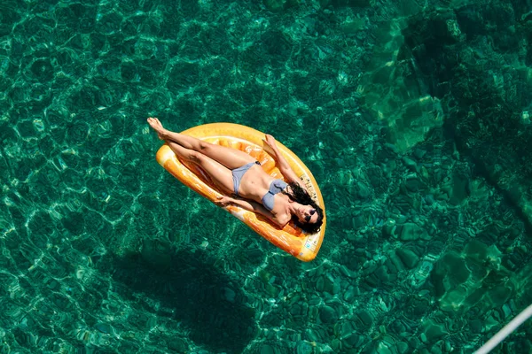 Bikinili sıska genç bir kadın ve şişme yatakta güneş gözlüğü açık denizde yüzüyor..