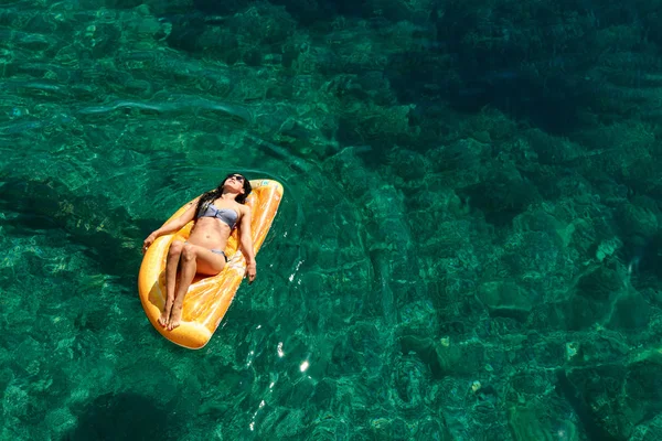 Bikinili sıska genç bir kadın ve şişme yatakta güneş gözlüğü açık denizde yüzüyor..