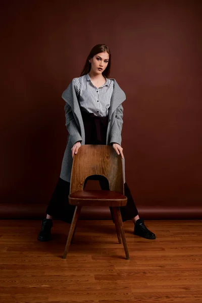 Independent young woman posing in the photostudio. Gorgeous young female in classic suit and grey coat. Brown background. Vintage chair style. Woman dressed in Man clothes. Fashionable clothes.