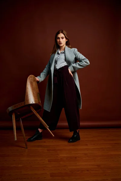 Independent young woman posing in the photostudio. Gorgeous young female in classic suit and grey coat. Brown background. Vintage chair style. Woman dressed in Man clothes. Fashionable clothes.