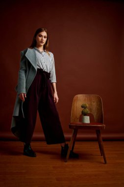 Fotostudio 'da poz veren bağımsız genç bir kadın. Klasik takım elbiseli ve gri ceketli güzel bir bayan. Kahverengi arka plan. Klasik sandalye stili. Erkek kıyafetleri giymiş bir kadın. Şık kıyafetler..
