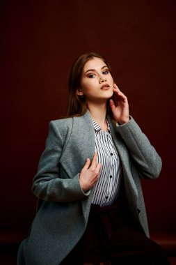 Fotostudio 'da poz veren bağımsız genç bir kadın. Klasik takım elbiseli ve gri ceketli güzel bir bayan. Kahverengi arka plan. Klasik sandalye stili. Erkek kıyafetleri giymiş bir kadın. Şık kıyafetler..