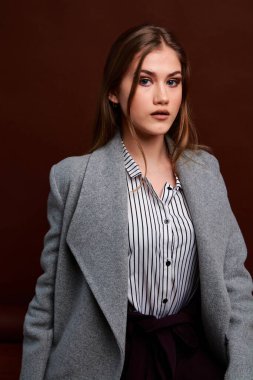 Fotostudio 'da poz veren bağımsız genç bir kadın. Klasik takım elbiseli ve gri ceketli güzel bir bayan. Kahverengi arka plan. Klasik sandalye stili. Erkek kıyafetleri giymiş bir kadın. Şık kıyafetler..