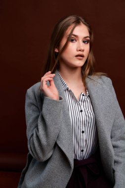 Fotostudio 'da poz veren bağımsız genç bir kadın. Klasik takım elbiseli ve gri ceketli güzel bir bayan. Kahverengi arka plan. Klasik sandalye stili. Erkek kıyafetleri giymiş bir kadın. Şık kıyafetler..