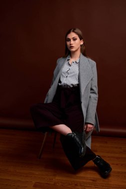 Fotostudio 'da poz veren bağımsız genç bir kadın. Klasik takım elbiseli ve gri ceketli güzel bir bayan. Kahverengi arka plan. Klasik sandalye stili. Erkek kıyafetleri giymiş bir kadın. Şık kıyafetler..