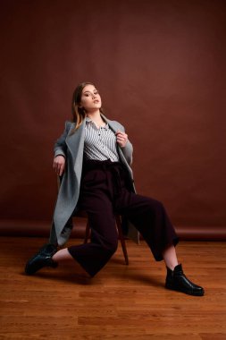 Fotostudio 'da poz veren bağımsız genç bir kadın. Klasik takım elbiseli ve gri ceketli güzel bir bayan. Kahverengi arka plan. Klasik sandalye stili. Erkek kıyafetleri giymiş bir kadın. Şık kıyafetler..