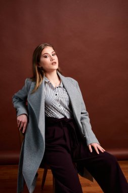 Fotostudio 'da poz veren bağımsız genç bir kadın. Klasik takım elbiseli ve gri ceketli güzel bir bayan. Kahverengi arka plan. Klasik sandalye stili. Erkek kıyafetleri giymiş bir kadın. Şık kıyafetler..