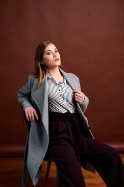 Fotostudio 'da poz veren bağımsız genç bir kadın. Klasik takım elbiseli ve gri ceketli güzel bir bayan. Kahverengi arka plan. Klasik sandalye stili. Erkek kıyafetleri giymiş bir kadın. Şık kıyafetler..
