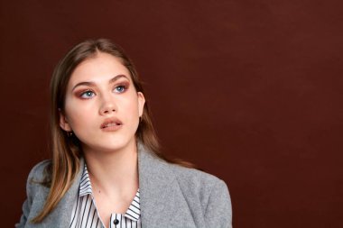 Fotostudio 'da poz veren bağımsız genç bir kadın. Klasik takım elbiseli ve gri ceketli güzel bir bayan. Kahverengi arka plan. Klasik sandalye stili. Erkek kıyafetleri giymiş bir kadın. Şık kıyafetler..