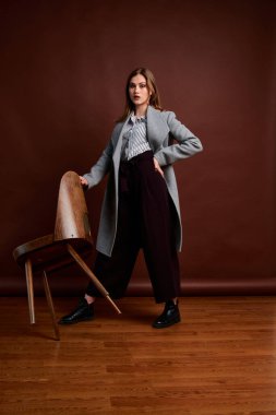 Fotostudio 'da poz veren bağımsız genç bir kadın. Klasik takım elbiseli ve gri ceketli güzel bir bayan. Kahverengi arka plan. Klasik sandalye stili. Erkek kıyafetleri giymiş bir kadın. Şık kıyafetler..
