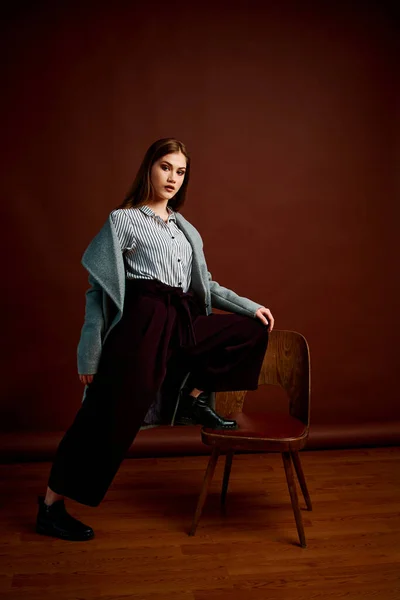 Independent young woman posing in the photostudio. Gorgeous young female in classic suit and grey coat. Brown background. Vintage chair style. Woman dressed in Man clothes. Fashionable clothes.
