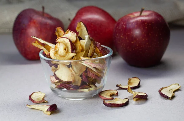 Dried Apple chips in a glass bowl and three red apples on a gray and linen background. Organic natural food. Close up. Side view.