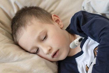 Pijamalı hasta çocuk dijital termometreyle yatakta yatıyor.