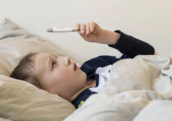 Vorschulkranker Junge im Pyjama liegt im Bett und schaut auf Thermometer — Stockfoto