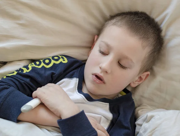 Niño enfermo preescolar en pijama acostado en la cama con un termómetro digital — Foto de Stock