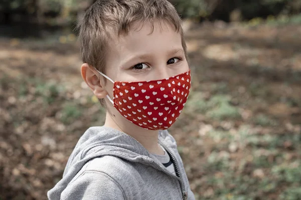 stock image Portrait of young boy wearing handmade cotton fabric face mask. Child in red mask with hearts in the park in sunny day. Protection against saliva, cough, dust, pollution, virus, bacteria, coronavirus, COVID-19. 