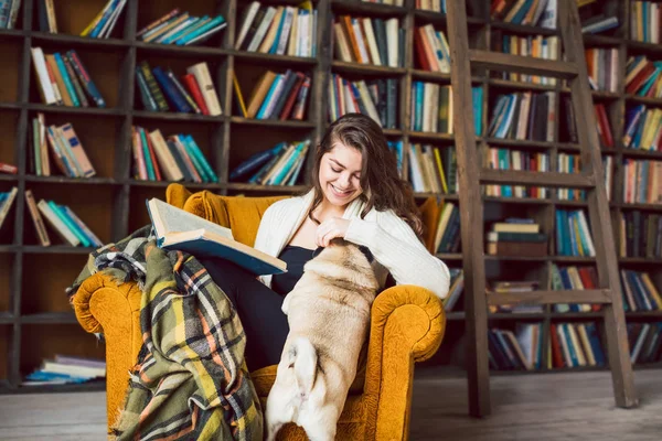 Fille assise sur la chaise dans la bibliothèque de la maison et jouer avec chien chiot gros chiot mignon — Photo