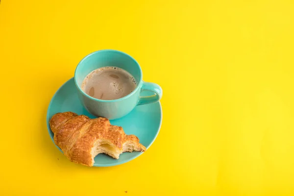 Croissant morso è su piatto blu e una tazza di cacao caffè sono su una tovaglia di sfondo giallo allegro. Un umore festoso e giocoso. Dolci da dessert croissant a colazione — Foto Stock