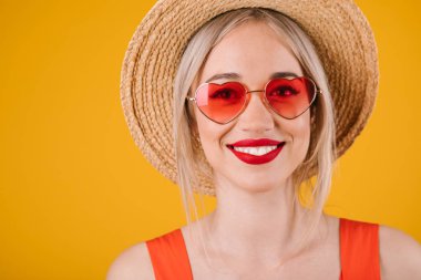 Sarı arka plan üzerinde şapkalı sarışın kadın. Kalp şeklinde pembe güneş gözlüğü. Sıcak yaz renkleri