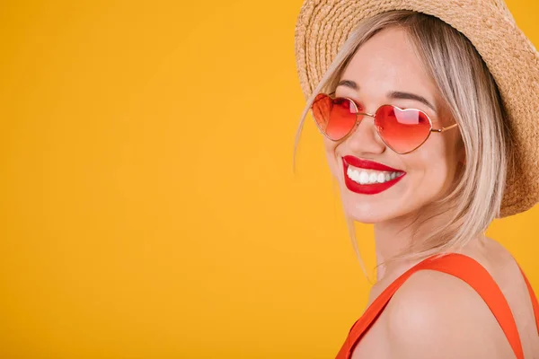Adorable mujer rubia con grandes dientes sonrisa en sombrero de paja sobre fondo amarillo brillante. Gafas de sol rosa en forma de corazón —  Fotos de Stock